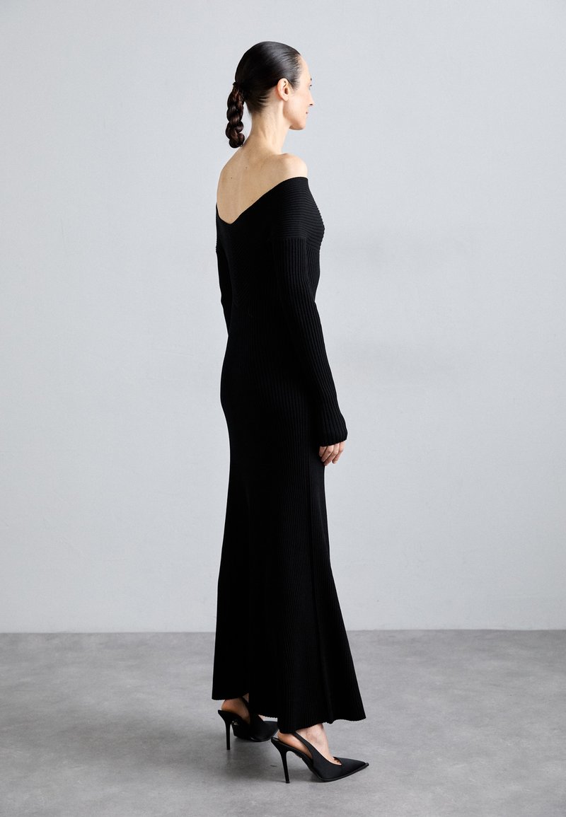 Robe noire côtelée décolletée, à manches longues, avec un corsage ajusté et un ourlet évasé, portée avec des chaussures à talons hauts noires. Texture lisse et design minimal.