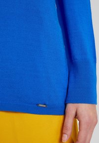 Sininen neulepaita pitkilla hihoilla ja ribbaiduilla hihansuilla; siinä on pieni metallilogo helman kohdalla. Paita on päälle puettuna keltaisen kankaan päällä.