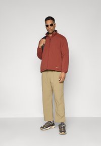 Veste isolante rouge avec un col haut, fermeture éclair à l'avant et un accent noir. Pantalons beige à jambes larges et chaussures de plein air marron avec des détails gris.