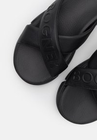 Sandales à enfiler en cuir noir avec des brides croisées embossées du nom de la marque, texture lisse et design à bout arrondi.