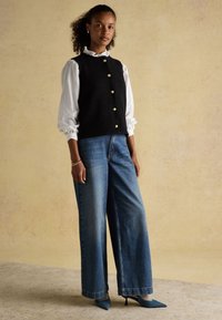 Woman standing wearing a white long-sleeve blouse, black buttoned vest, wide-leg blue jeans, and teal pointed heels against a beige background.