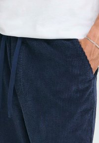 Personne portant un pantalon en velours côtelé bleu marine avec une main dans la poche et un bracelet en argent au poignet, associé à un haut blanc.