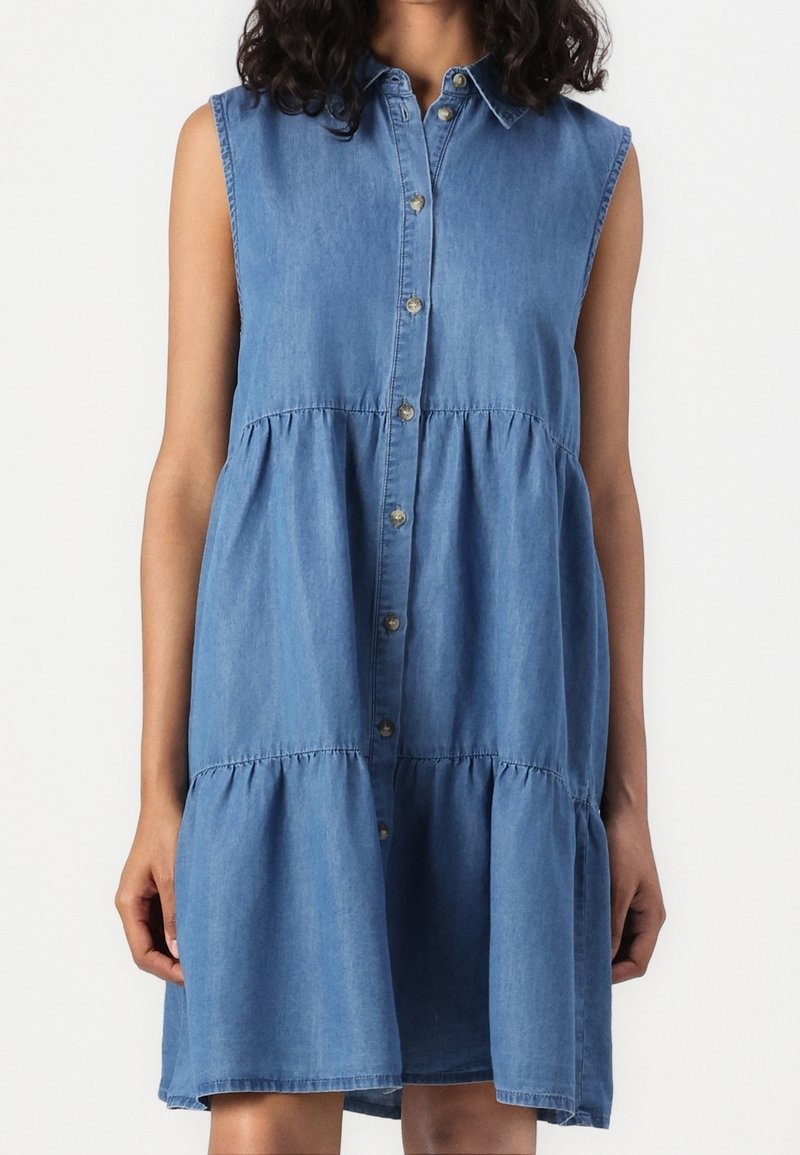Robe en jean bleue sans manches à boutons avec une jupe à volants, portée par une personne aux cheveux bouclés foncés sur un fond clair uni.