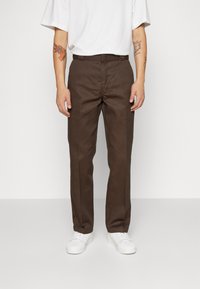 Man som bär bruna skräddarsydda byxor, vita sneakers, och en vit t-shirt med tatueringar synliga på båda underarmarna.