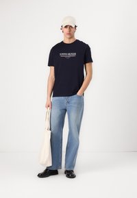 Marineblå T-shirt med hvid "TOMMY HILFIGER" logo, båret med lyseblå jeans og sorte sko. En beige tote taske holdes i den ene hånd.