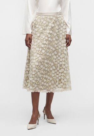 Jupe midi avec broderie florale crème sur tissu beige, portée avec un chemisier blanc et des chaussures à talons crème.