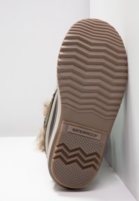 Sohle eines Winterstiefels mit braunem Gummiprofil und dem Aufkleber "WASSERDICHT", der Stiefel lehnt an der Ecke zwischen weißer Wand und Boden.