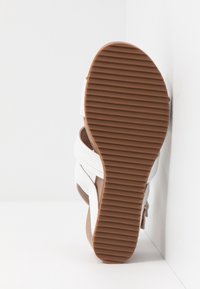 Sandale blanche avec une semelle en caoutchouc marron, présentant une surface texturée et des rainures horizontales. Brides ajustables pour un maintien sécurisé.