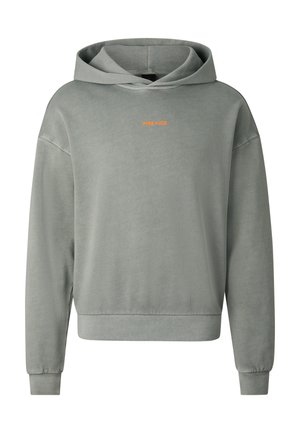 Jonge man die een lichtgrijze hoodie met capuchon draagt, bijpassende korte broek, witte sokken, grove sneakers en grote geel getinte zonnebril, staand.