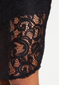 Robe en dentelle noire avec des motifs en spirales complexes, des panneaux semi-transparents et un ourlet festonné. Texture douce avec des détails délicats.