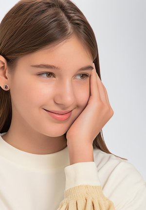 Nahaufnahme einer jungen Person mit hellbraunem Haar, die einen cremefarbenen Pullover mit gepufften Ärmeln und Ohrsteckern mit roten und schwarzen Akzenten trägt.