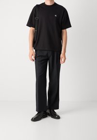 Czarna bawełniana koszulka z okrągłym dekoltem i detalem logo, zestawiona z czarnymi spodniami i sznurowanym obuwiem, stojąca na neutralnym tle.