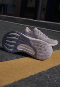 Zapatillas de correr lilas con un empeine texturizado, diseño aerodinámico y suelas de goma con ranuras y crestas. Colocadas sobre asfalto junto a una línea amarilla.