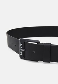 Ceinture en cuir noir avec une finition lisse, boucle en métal rectangulaire avec un logo et plusieurs trous de réglage.