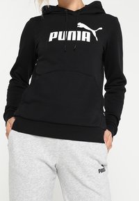 Sweat à capuche noir avec logo "PUMA" blanc, poche avant, manches longues et coupe décontractée. Associé à un pantalon de survêtement gris avec logo.