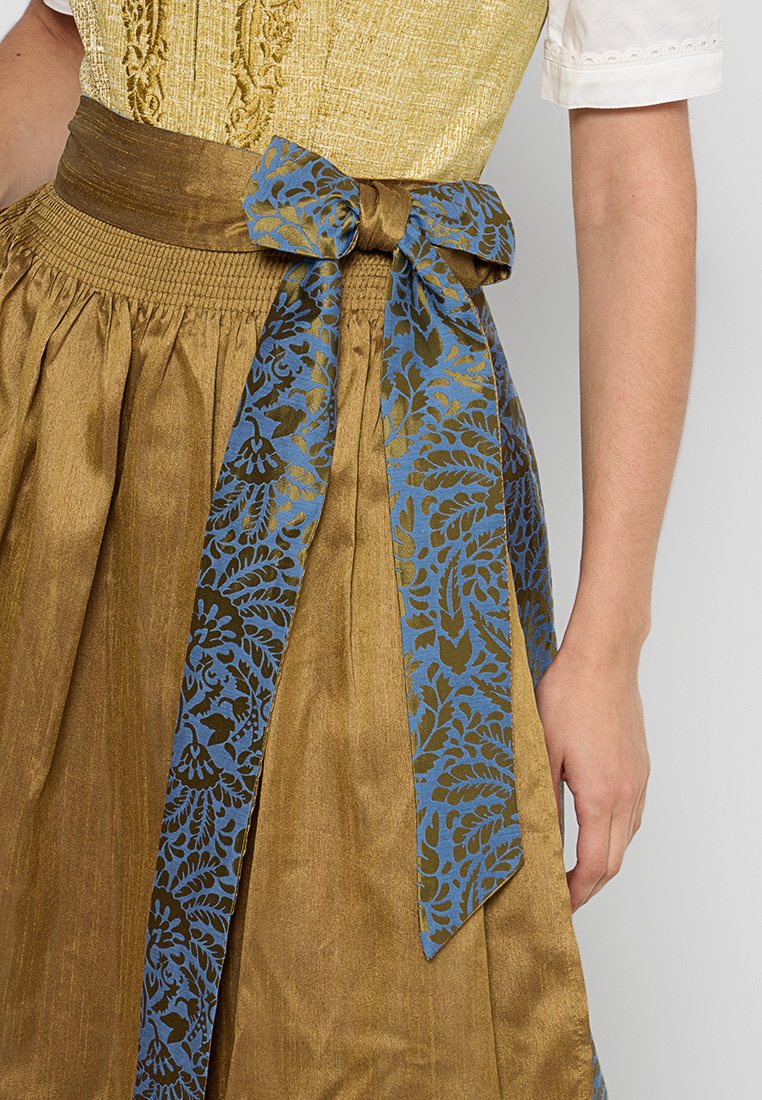 Goldener Rock mit gerafftem Bund, gebundenem braunen Gürtel mit einer blau gemusterten Schleife, getragen mit einem strukturierten gelben Oberteil und einer weißen Bluse mit kurzen Ärmeln.