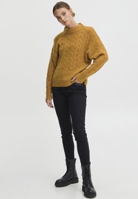 Senfgelber, gestrickter Pullover mit strukturiertem Muster, geripptem Saum und langen Ärmeln, kombiniert mit schwarzen Slim-Fit-Jeans und klobigen schwarzen Stiefeln.