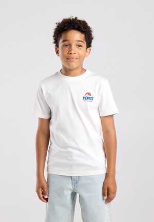 Jongen met krullend haar die een wit T-shirt met de tekst "Venice" en lichtblauwe spijkerbroek draagt, staand tegen een effen lichte achtergrond.