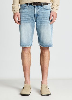 Uomo con pantaloncini in denim azzurro chiaro, maglione beige, cintura nera e scarpe beige senza lacci, in piedi davanti a uno sfondo semplice.