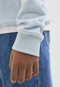 Felpa a manica lunga di colore azzurro chiaro con polsini a coste, abbinata a jeans scuri. Primo piano del tessuto testurizzato e del dettaglio della mano.