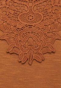 Tecido bronze com delicados detalhes em renda na parte superior, apresentando padrões florais e bordas texturizadas para um interesse visual adicional.