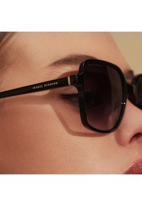 Lunettes de soleil carrées noires avec des verres foncés, présentant des accents dorés sur les branches et la marque "ISABEL BERNARD" sur le côté.