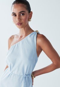 Světle modré šaty s asymetrickým výstřihem, texturovanou látkou a ozdobnými kovovými doplňky na rameni. Zavazují se v pase. Zlaté náušnice.