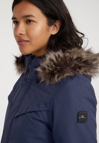 Marineblauwe parka-jack met faux fur-afwerking op de capuchon, voorzien van een ritsluiting, zijzakken en een merkiem op de mouw.
