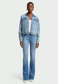 Woman wearing light blue denim jacket and high-waist blue flare jeans with white pointed shoes, standing against plain background.