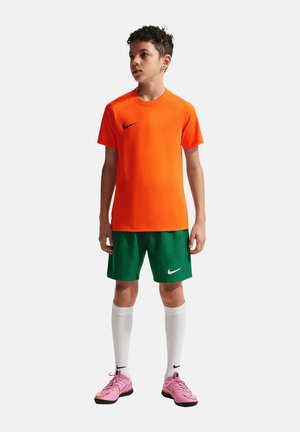 Junge trägt ein orangefarbenes Nike-Shirt, grüne Shorts, weiße knielange Socken und pinke Nike-Fußballschuhe, steht und schaut zur Seite.
