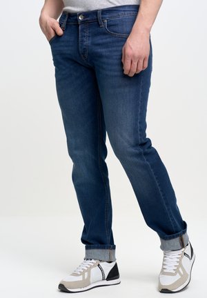 Vaqueros de mezclilla azul oscuro con corte recto, cinco bolsillos y un puño de color azul claro. Combinados con zapatillas grises y negras.