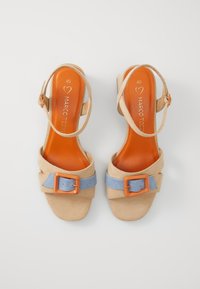 Sandales en daim beige avec une large bride bleue, un détail de boucle orange, et une bride de cheville. Semelle intérieure lisse de couleur orange vif.