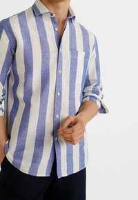 Homme portant une chemise à rayures verticales bleues et blanches à manches longues, une manche retroussée, et un pantalon noir.