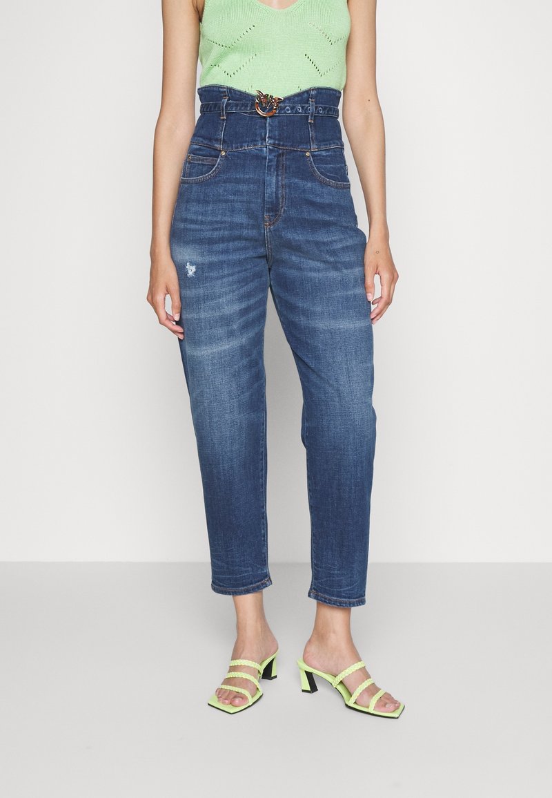 Femme portant un jean bleu taille haute avec une ceinture, un top sans manches vert et des sandales à talons vert citron, debout devant un fond blanc.