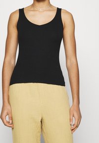 Débardeur noir à côtes avec un décolleté en V et des bretelles larges, associé à un pantalon texturé jaune clair. Le tissu semble doux et ajusté.