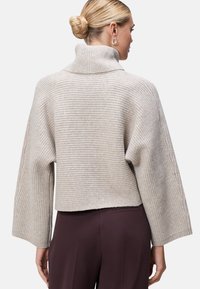 Hellbeige gestrickter Pullover mit hohem Kragen, weiten Ärmeln, gerippter Textur und cropped Design, kombiniert mit dunkelburgunderfarbenen Hosen.