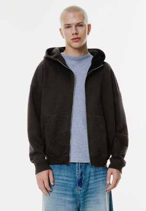 Dark brown zip-up hoodie with two front pockets, worn over a light gray t-shirt. Accompanied by light blue, faded denim shorts.