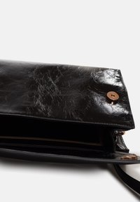 Borsa a tracolla in pelle nera, texture opaca, con chiusura a pattina e bottone a pressione color rame. L'interno è parzialmente visibile. Nessun motivo esterno visibile.