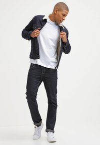 Mørk denimjakke med hvid shearlingkrave, hvid t-shirt, mørke jeans og hvide sneakers; modellen poserer og justerer jakken.