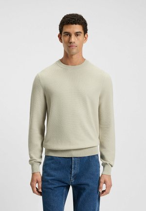 Light beige knitted sweater with round neckline and long sleeves. Features ribbed cuffs and hem. Paired with blue denim jeans.