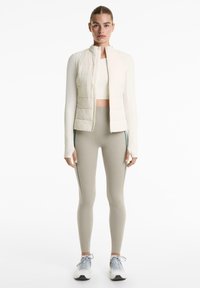 Femme se tenant de face, portant un haut blanc à manches longues raccourci, un gilet matelassé crème, des leggings beige clair avec des rayures sur les côtés, et des baskets grises.
