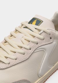 Κοντινή λήψη μπεζ αθλητικού παπουτσιού με υφή δέρματος, λεπτομέρειες από σουέντ, χοντρές κρεμ κορδόνια και ετικέτα με λογότυπο στη μαξιλαρωμένη γλώσσα.