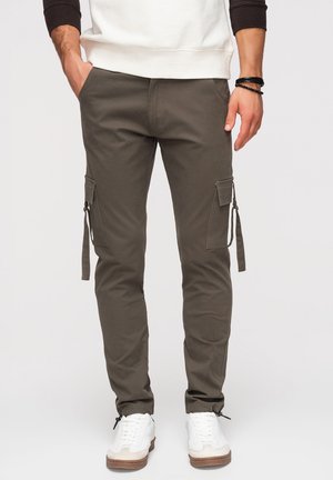 Mężczyzna noszący oliwkowe spodnie typu cargo z bocznymi kieszeniami i białe trampki, stoi z częściowo wsadzonymi w kieszenie rękami.