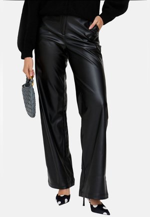 Frau trägt schwarze Lederhose mit weitem Bein, schwarzen Pullover, gepunktete High Heels und hält eine kleine graue gewebte Handtasche.