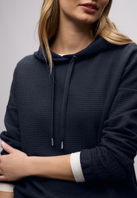 Gros plan sur une personne portant un sweat à capuche texturé foncé par-dessus une chemise blanche à manches longues, les bras croisés, les lèvres légèrement entrouvertes.