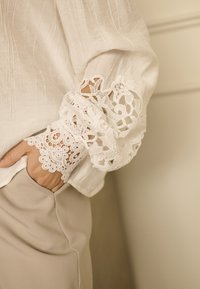 Blusa bianca con tessuto testurizzato e maniche rifinite in pizzo. Il pizzo presenta motivi floreali intricati, abbinati a pantaloni beigi aderenti.