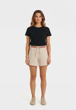 Donna che indossa una t-shirt nera a maniche corte, pantaloncini beige a costine con vita con coulisse e sneaker beige, in piedi davanti a uno sfondo bianco.