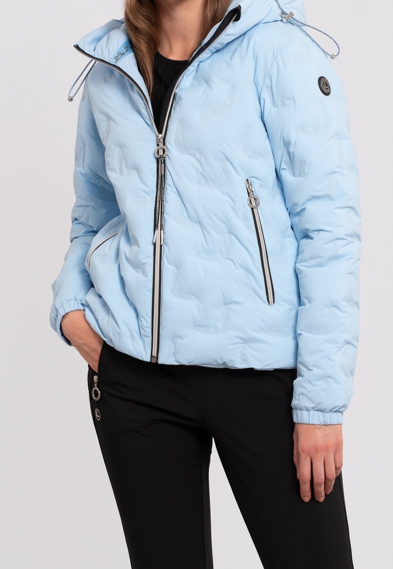 Femme portant une veste matelassée bleu clair avec fermeture éclair à l'avant et poches zippées, associée à un pantalon noir avec détails zippés.