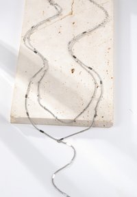 Zilveren ketting met meerdere strengen, met verschillende texturen en kleine kralen, gelegd op een lichtgekleurde, gestructureerde stenen ondergrond.