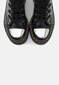 Botas de cuero negras con punteras plateadas, cordones negros y costuras amarillas. Tienen una punta redondeada y suela de goma texturizada.